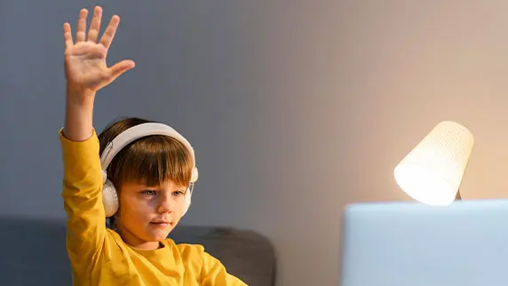 Picture of a child sitting at a desk with a laptop and headphones on while raising his hand