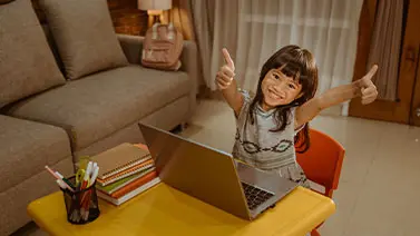 Picture of a child sitting at a desktop with a laptop on it smiling and giving two thumbs up