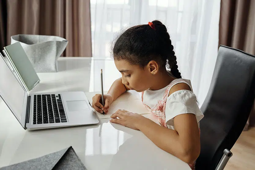 Child at desk with computer open, writing on paper