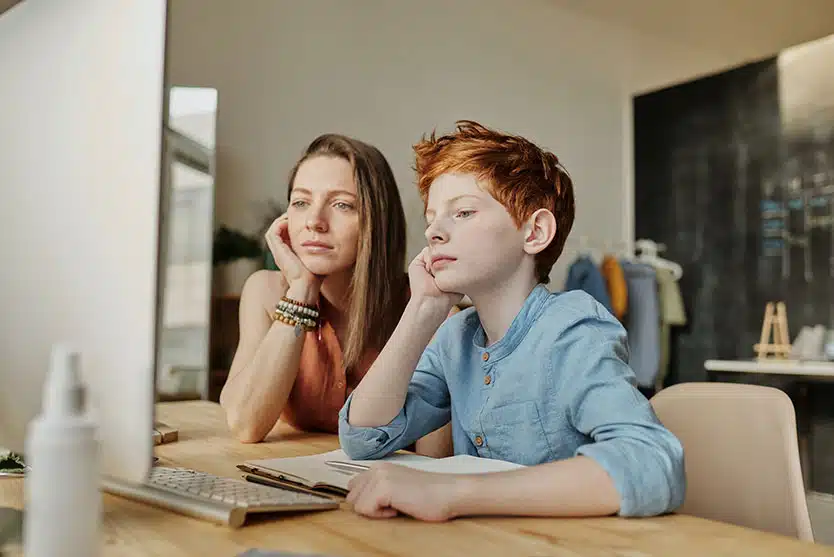 A mother and son sit at a desk in front of a computer