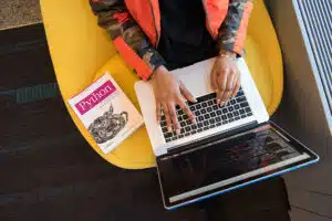 woman types on laptop seen vertically from above next to a Python textbook