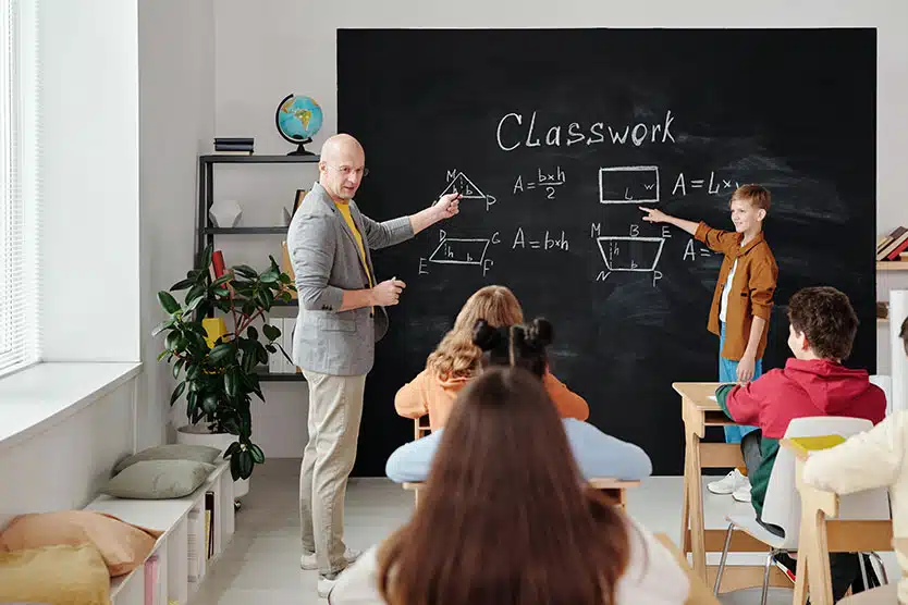 teacher and young student stand in front of class, writing math problems on blackboard