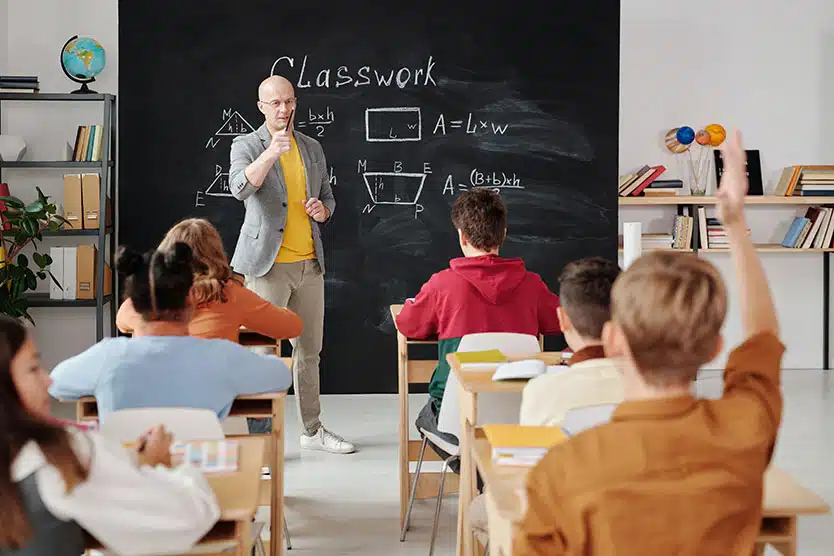 teacher in front of blackboard, students in front, one with raised hand