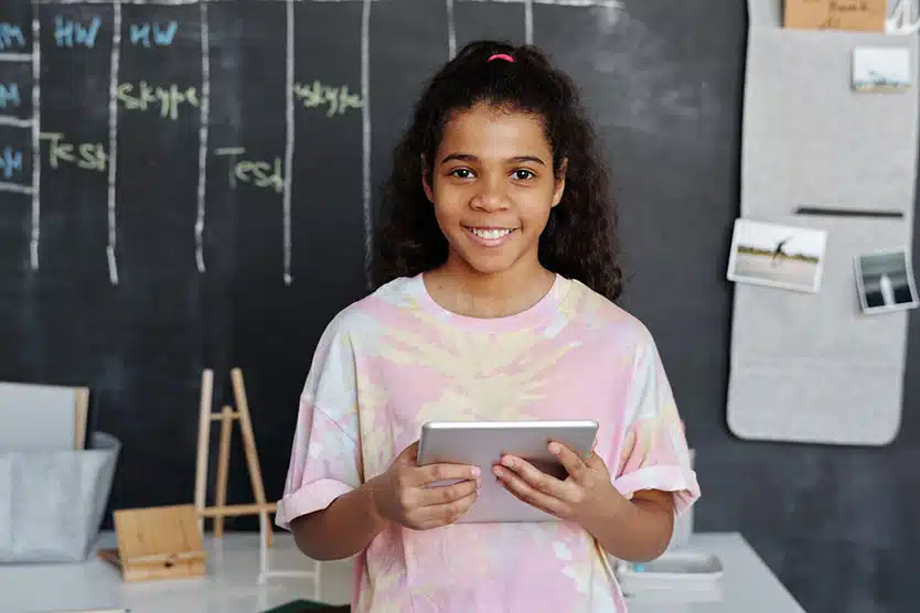 A smiling little girl wearing a pink tie-dye t-shirt, holding a silver tablet. She’s standing in front of a chalkboard