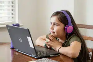 A little girl with long brown hair sat with her hands clasped, watching a laptop. She’s wearing big purple and pink headphones