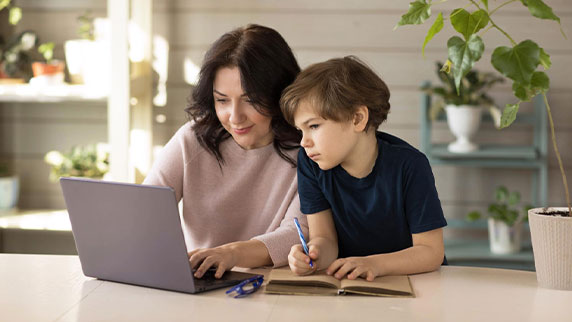 Mother and son engaged in online classes sitting infront of a laptop and making notes in a notebook