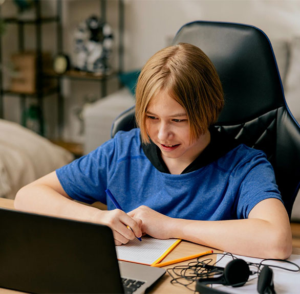 Boy sitting at desk with pen in hand writing notes looking at laptop