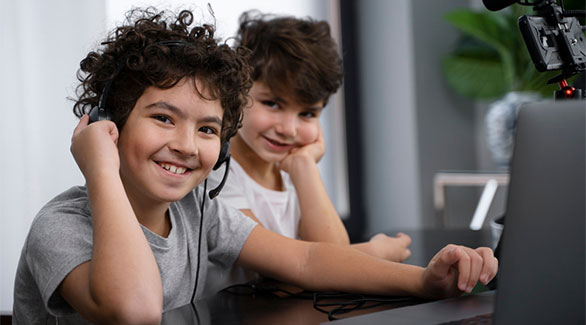 Two young kids sitting at desk learning online with laptop smiling at the camera