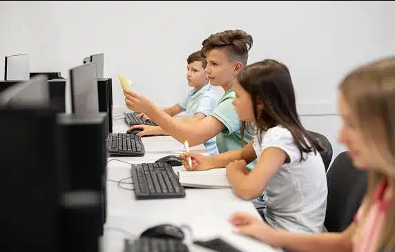 View from behind a child sitting at a desktop looking at a laptop with code on the screen